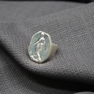 K&ouml;nnte beinhalten: Ein silberner Ring mit einer achteckigen Fl&auml;che, die ein Reliefbild einer stehenden Person zeigt. Der Ring liegt auf einem grauen Stoffhintergrund.