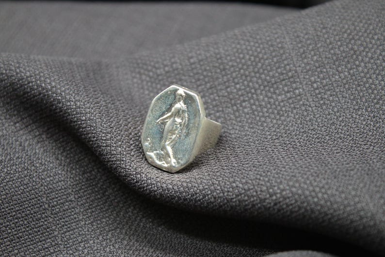 K&ouml;nnte beinhalten: Ein silberner Ring mit einer achteckigen Fl&auml;che, die ein Reliefbild einer stehenden Person zeigt. Der Ring liegt auf einem grauen Stoffhintergrund.