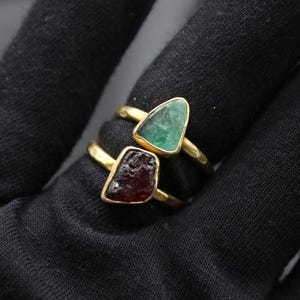May include: Two gold rings with green and red gemstone settings. The rings are on a black fabric background.