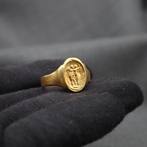May include: A gold ring with an oval-shaped design featuring two figures holding hands. The ring is set against a dark gray background.