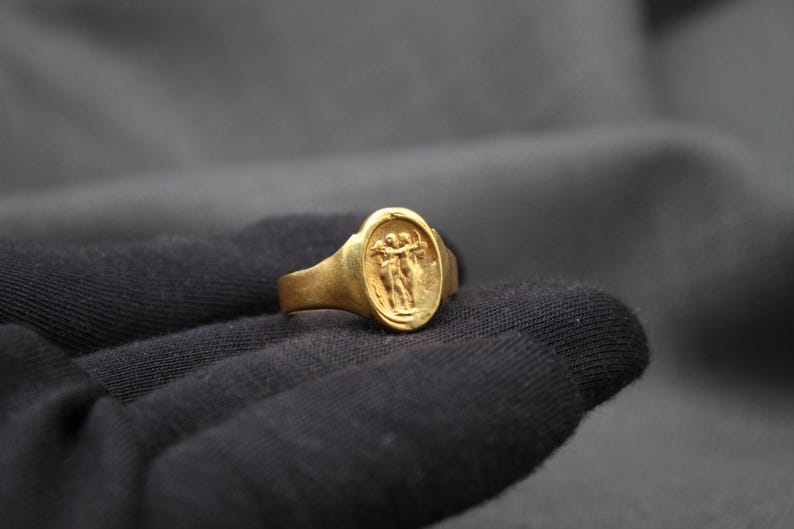 May include: A gold ring with an oval-shaped design featuring two figures holding hands. The ring is set against a dark gray background.