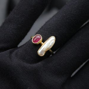 Puede incluir: Un anillo de oro con una piedra preciosa rosa y una perla blanca. El anillo est&aacute; sobre una superficie negra.