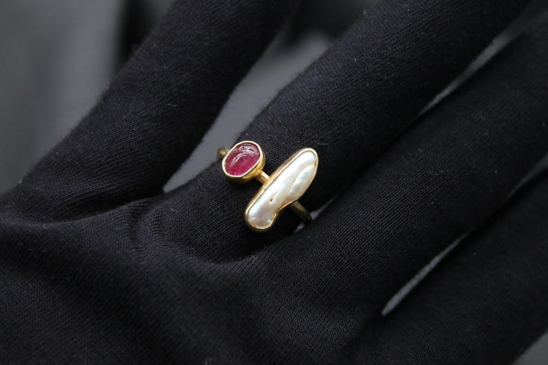 Puede incluir: Un anillo de oro con una piedra preciosa rosa y una perla blanca. El anillo est&aacute; sobre una superficie negra.