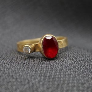 May include: A gold ring featuring a red oval gemstone and a smaller clear gemstone. The ring has a hammered finish.