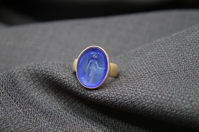 May include: A gold ring with a blue oval-shaped gemstone featuring a carved design of a person.