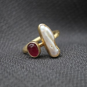 May include: Gold ring featuring a white pearl and a red gemstone. The ring is displayed against a grey fabric backdrop.