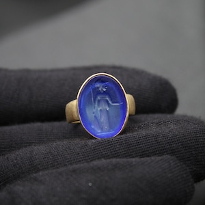 May include: A gold ring with a blue oval-shaped stone featuring a carved design of a woman holding a staff.