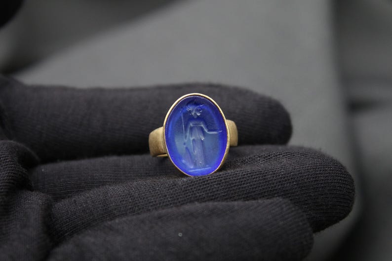 May include: A gold ring with a blue oval-shaped stone featuring a carved design of a woman holding a staff.