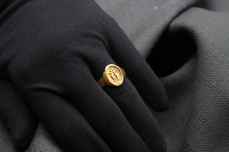 May include: A gold ring with a detailed design of two figures holding hands. The ring is worn on a person's finger.