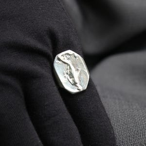 K&ouml;nnte beinhalten: Ein silberner Ring mit einem detaillierten Design einer stilisierten Figur. Der Ring wird an einem Finger getragen.