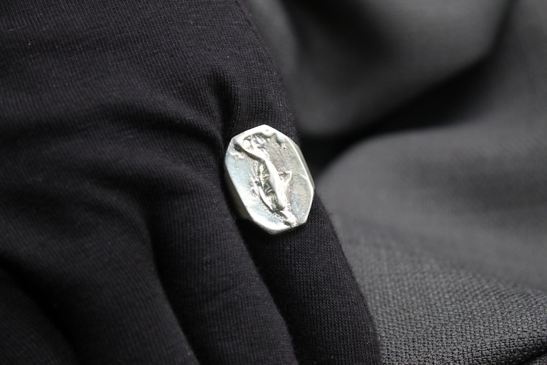 K&ouml;nnte beinhalten: Ein silberner Ring mit einem detaillierten Design einer stilisierten Figur. Der Ring wird an einem Finger getragen.