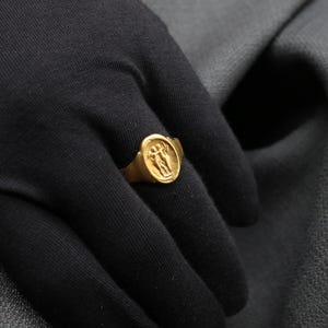May include: A gold ring with a raised design of two figures holding hands. The ring is worn on a person's finger.