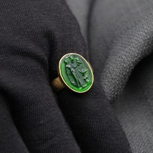 May include: A gold ring with a green oval-shaped stone featuring a carved design of a person holding a bird.