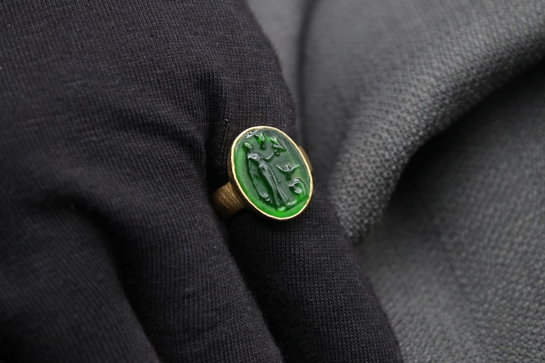 May include: A gold ring with a green oval-shaped stone featuring a carved design of a person holding a bird.