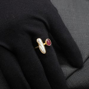 Puede incluir: Un anillo de oro con una piedra blanca nacarada y una peque&ntilde;a piedra roja. El anillo est&aacute; sobre un fondo de tela negra.