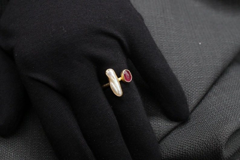 Puede incluir: Un anillo de oro con una piedra blanca nacarada y una peque&ntilde;a piedra roja. El anillo est&aacute; sobre un fondo de tela negra.