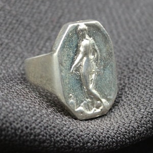 K&ouml;nnte beinhalten: Ein silberner Ring mit einer achteckigen Fl&auml;che, die ein erhabenes Design einer stehenden Person mit herabh&auml;ngenden Armen zeigt.