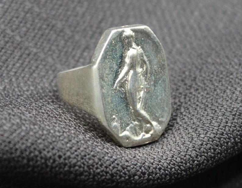 K&ouml;nnte beinhalten: Ein silberner Ring mit einer achteckigen Fl&auml;che, die ein erhabenes Design einer stehenden Person mit herabh&auml;ngenden Armen zeigt.