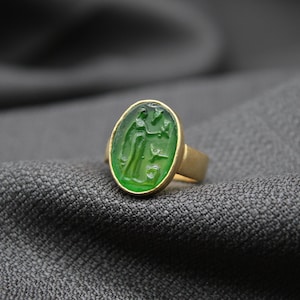May include: A gold ring with a green oval stone featuring a carved design. The ring is on a gray fabric background.