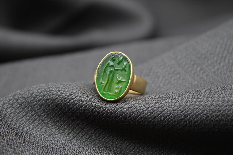 May include: A gold ring with a green oval stone featuring a carved design. The ring is on a gray fabric background.