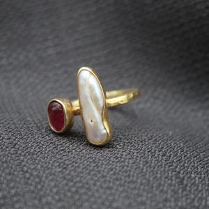 Puede incluir: Anillo de oro con una perla blanca y una piedra roja. El anillo tiene una banda texturizada.