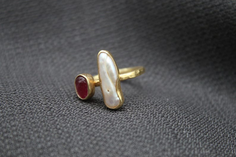 Puede incluir: Anillo de oro con una perla blanca y una piedra roja. El anillo tiene una banda texturizada.