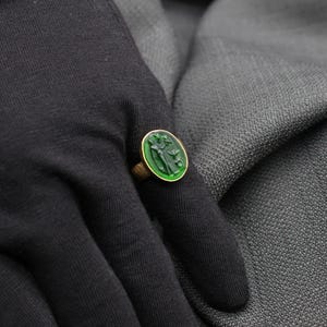 May include: A gold ring with a green gemstone featuring a carved design. The ring is worn on a finger.