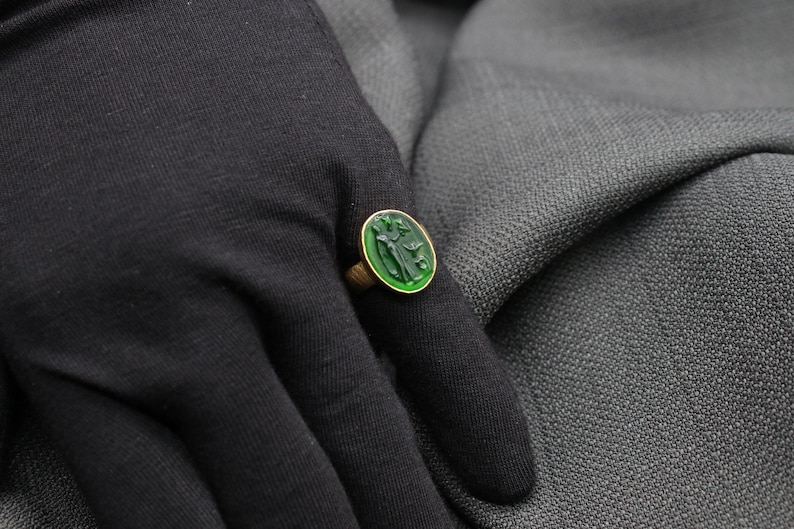 May include: A gold ring with a green gemstone featuring a carved design. The ring is worn on a finger.