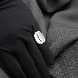 K&ouml;nnte beinhalten: Ein silberner Ring mit einem strukturierten Design, das einen stilisierten Vogel zeigt. Der Ring wird an einem Finger getragen, der mit schwarzem Stoff bedeckt ist.