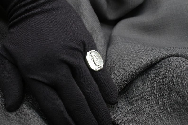 K&ouml;nnte beinhalten: Ein silberner Ring mit einem strukturierten Design, das einen stilisierten Vogel zeigt. Der Ring wird an einem Finger getragen, der mit schwarzem Stoff bedeckt ist.