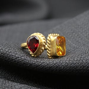 May include: A gold ring with two gemstones. The ring has a red heart-shaped gemstone on one side and a yellow rectangular gemstone on the other side.