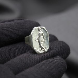 K&ouml;nnte beinhalten: Ein silberner Ring mit einem erhabenen Design einer Frau, die auf einem Sockel steht. Der Ring ist oval geformt und hat eine glatte, polierte Oberfl&auml;che.