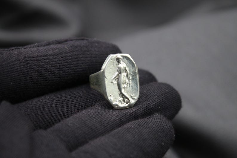 K&ouml;nnte beinhalten: Ein silberner Ring mit einem erhabenen Design einer Frau, die auf einem Sockel steht. Der Ring ist oval geformt und hat eine glatte, polierte Oberfl&auml;che.