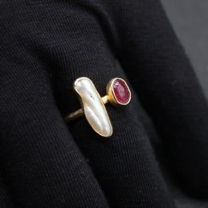 Puede incluir: Un anillo de oro con una piedra blanca nacarada y una piedra roja. El anillo est&aacute; sobre un fondo de tela negra.