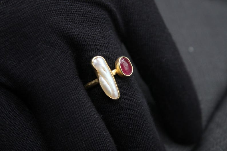 Puede incluir: Un anillo de oro con una piedra blanca nacarada y una piedra roja. El anillo est&aacute; sobre un fondo de tela negra.