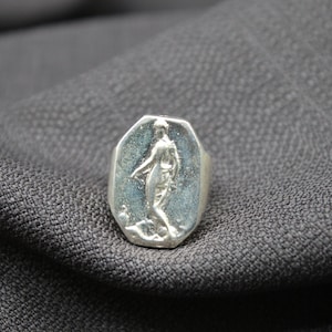 K&ouml;nnte beinhalten: Ein silberner Ring mit einem ovalen Design, das ein erhabenes Bild einer Person zeigt. Der Ring liegt auf einem grauen Stoffhintergrund.