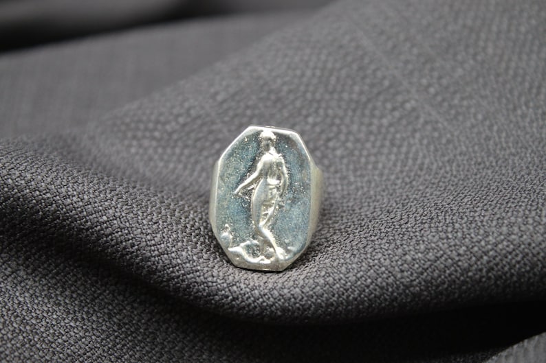 K&ouml;nnte beinhalten: Ein silberner Ring mit einem ovalen Design, das ein erhabenes Bild einer Person zeigt. Der Ring liegt auf einem grauen Stoffhintergrund.