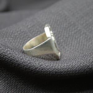K&ouml;nnte beinhalten: Ein silberner Ring mit einem detaillierten Design auf der Oberseite. Der Ring liegt auf einem grauen Stoffhintergrund.