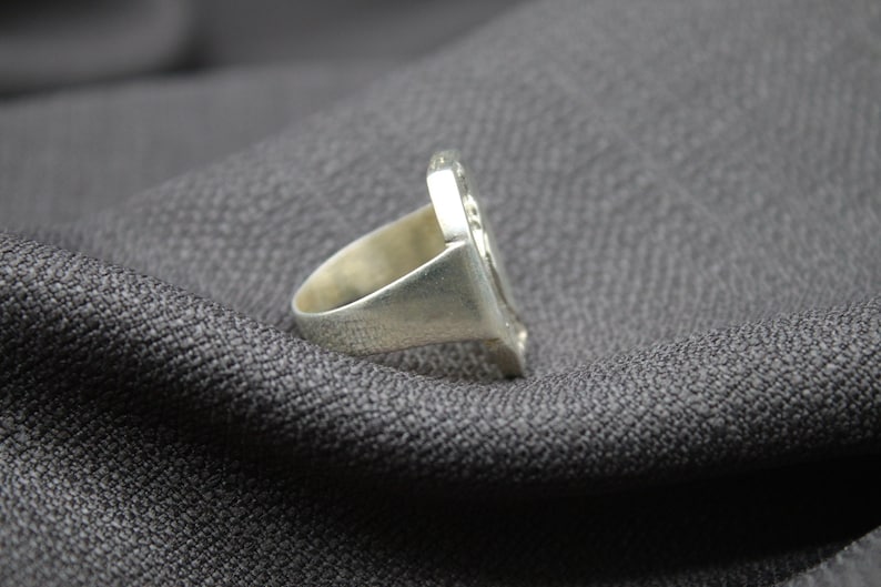 K&ouml;nnte beinhalten: Ein silberner Ring mit einem detaillierten Design auf der Oberseite. Der Ring liegt auf einem grauen Stoffhintergrund.