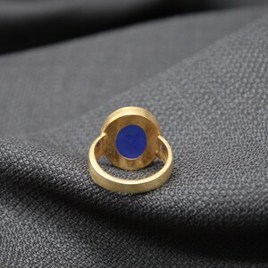 May include: A gold ring with a blue oval gemstone set in the center. The ring is on a gray fabric background.