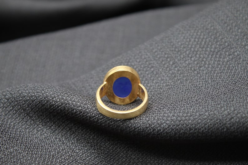 May include: A gold ring with a blue oval gemstone set in the center. The ring is on a gray fabric background.