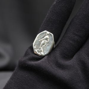 K&ouml;nnte beinhalten: Ein silberner Ring mit einem detaillierten Design einer stehenden Figur auf einer Basis. Der Ring liegt auf einem schwarzen Stoffhintergrund.