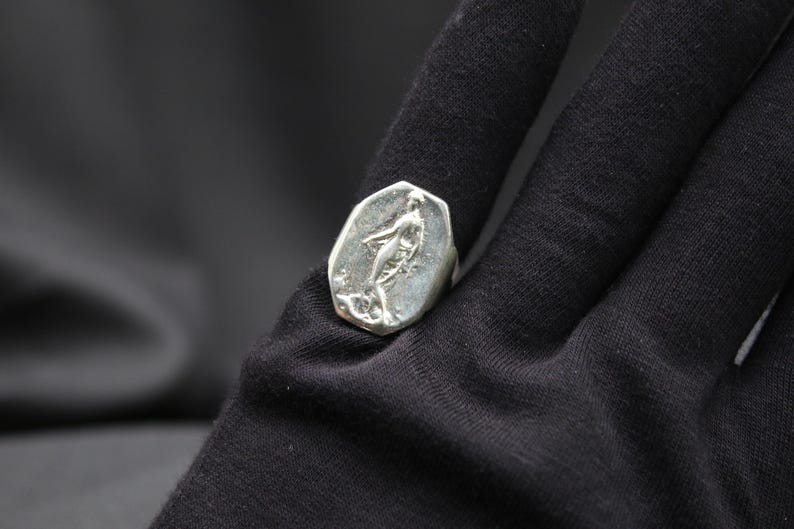 K&ouml;nnte beinhalten: Ein silberner Ring mit einem detaillierten Design einer stehenden Figur auf einer Basis. Der Ring liegt auf einem schwarzen Stoffhintergrund.
