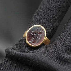May include: A gold ring with a dark red oval gemstone featuring a carved design. The ring is worn on a finger.
