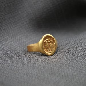 May include: A gold ring with an oval-shaped setting featuring a raised design of two figures holding hands.