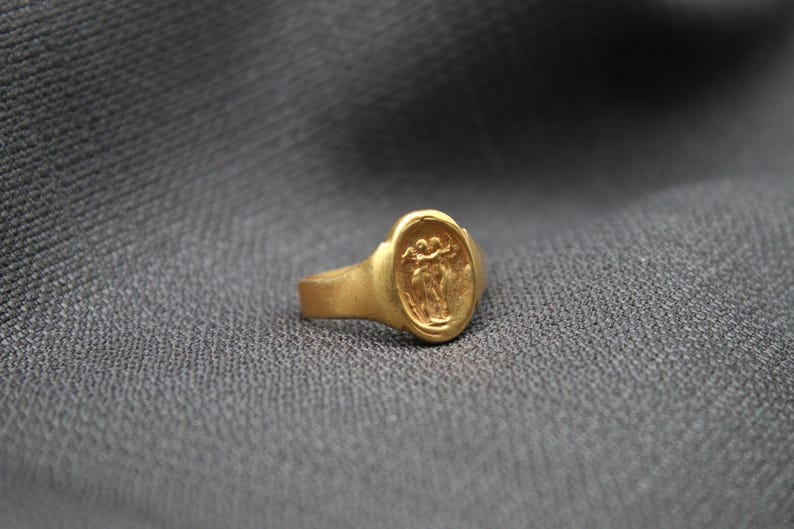 May include: A gold ring with an oval-shaped setting featuring a raised design of two figures holding hands.