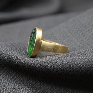 May include: A gold ring with a green oval gemstone. The ring is set on a gray fabric background.