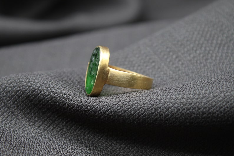 May include: A gold ring with a green oval gemstone. The ring is set on a gray fabric background.