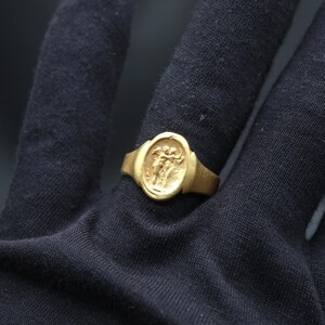May include: A gold ring with an oval-shaped bezel featuring a raised design of two figures. The ring is worn on a black fabric surface.