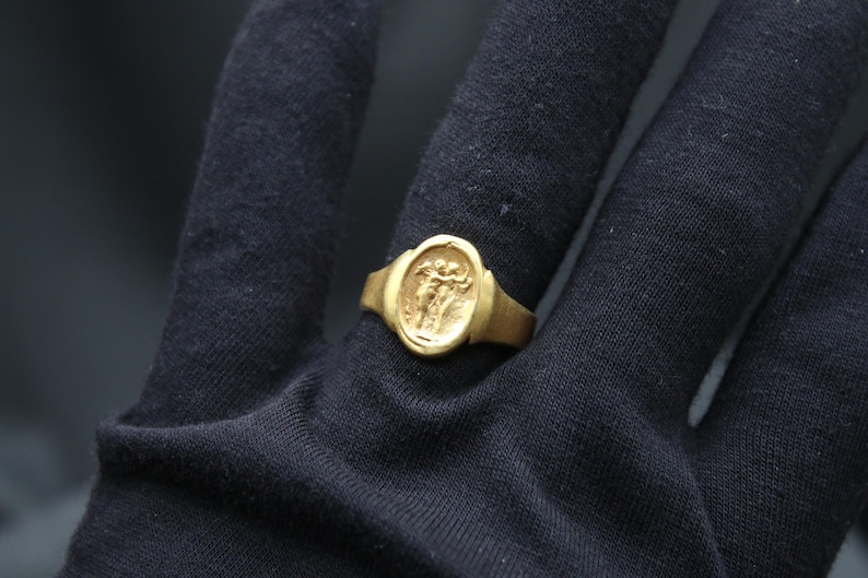 May include: A gold ring with an oval-shaped bezel featuring a raised design of two figures. The ring is worn on a black fabric surface.